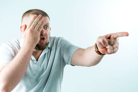 An angry bearded man in a white T-shirt on a white background closed one eye with his hand and pointed at someone with the other hand. Hes kind of angry.の写真素材