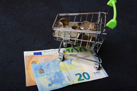 Shopping cart full of coins and banknotes on dark backgroundの写真素材