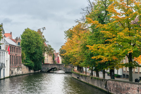 A picturesque canal in Bruges, Belgium, featuring an old stone bridge and autumn trees with orange leaves reflecting in the water, capturing the charm of the historic city.の写真素材
