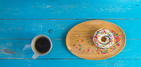 Donut with white cream topped with chocolate and sprinkle with sugar in a variety of colors in a wooden tray, and a cup of black coffee, Placed on a blue vintage wooden table.の写真素材