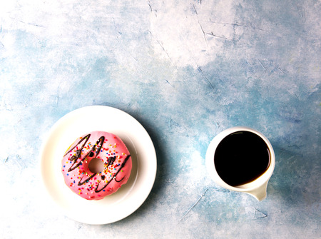 Donut coated with strawberry cream. Topped with chocolate and colors sugar in a white ceramic dish. With black coffee cup.の写真素材