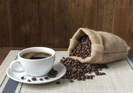Black coffee in ceramic cup with saucer Double sack with roasted coffee beans. Placed on a light brown tablecloth with blue patterns. The background is a vintage wooden pattern.の写真素材