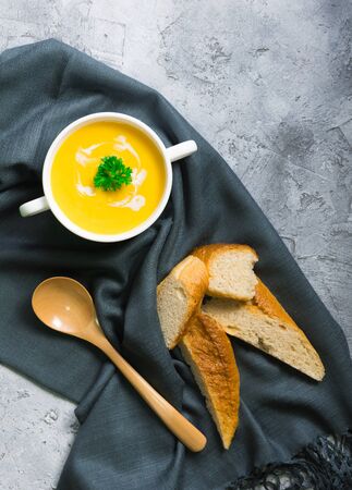 Pumpkin soup packed in a white ceramic cup. Serve with baguettes placed on a dark gray tablecloth along with a wooden spoon. Everything is laid on a white uneven floor.の写真素材
