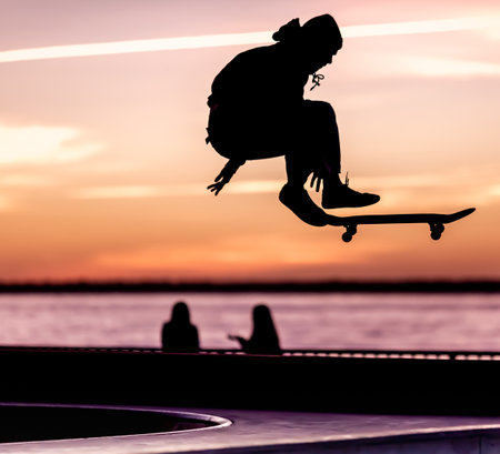 Unrecognizable teenage boy silhouette showing high jump tricks on scooter against orange sunsetwarm...
