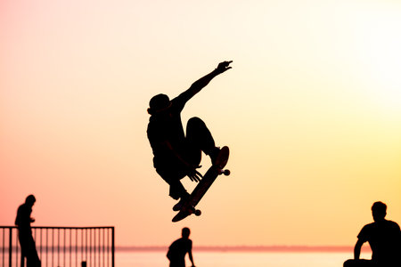 Unrecognizable teenage boy silhouette showing high jump tricks on scooter against orange sunsetwarm...