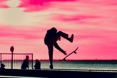Unrecognizable teenage boy silhouette showing high jump tricks on scooter against orange sunsetwarm sky at skatepark. Sport, extreme, freestyle, outdoor activity conceptの写真素材