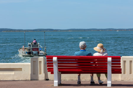 Rear view of an older couple sitting close together on a bench looking out over the water.の写真素材