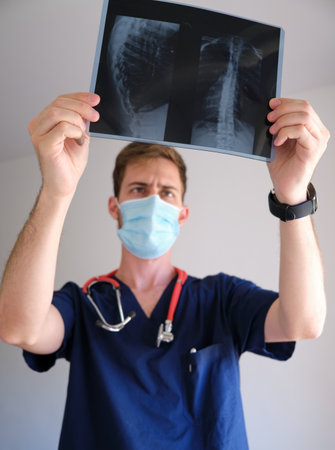 a handsome physician men wearing scrub, stethoscope and face mask looking to an x-ray during the sars cov 2 pandemicの写真素材