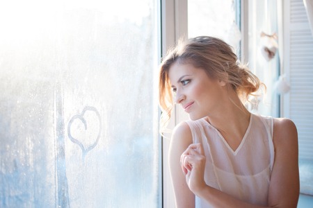 beautiful woman with fresh daily makeup and romantic wavy hairstyle, sitting on the windowsill and draws on glassの写真素材
