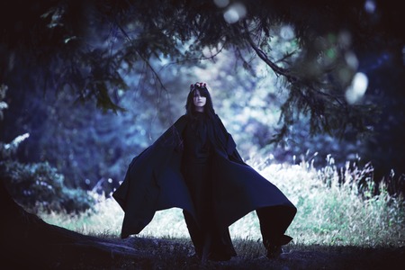 young black-haired girl in a long black cloak with a hood in a dark forestの写真素材