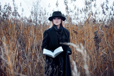 Portrait of young attractive woman in a black coat and hat. She's one in a field reading a book, autumn landscape, dry grassの写真素材