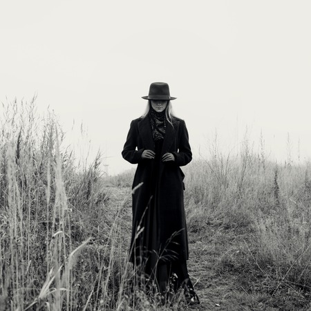 Portrait of young attractive woman in a black coat and hat. She's one in a field reading a book,...