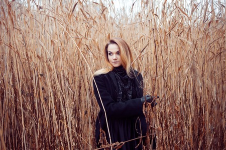 Portrait of young attractive woman in a black coat and hat. She's one in a field reading a book,...