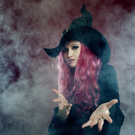 Attractive woman in witches hat and costume with red hair performs magic on a pink background. Halloween, horror theme.の写真素材