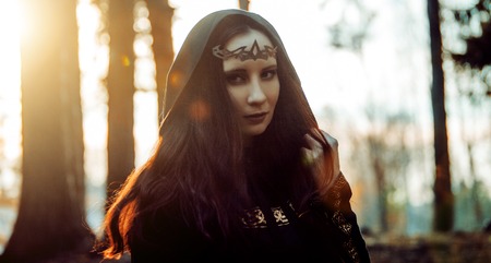 Young beautiful and mysterious woman in the woods, in a black cloak with a hood, the image of a forest elf or witchの写真素材