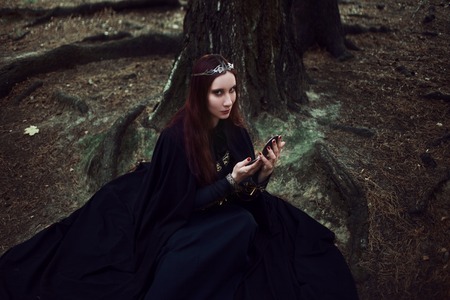 Young beautiful and mysterious woman in the woods, in a black cloak with a hood, the image of a forest elf or witchの写真素材