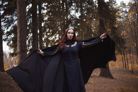 Young beautiful and mysterious woman in woods, in black cloak with hood, image of forest elf or witchの写真素材