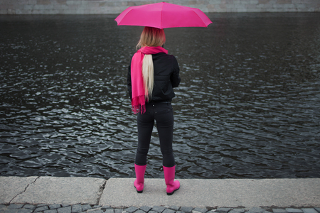 Girl in a bright pink scarf, rubber boots and umbrella standing on the banks of the river, back. Grey grim landscape.の写真素材