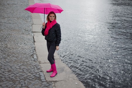 Beautiful young and happy blond woman in a bright pink scarf, rubber boots and umbrella walking in a rainy city.の写真素材