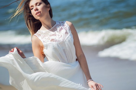 Young charming brunette woman on sea coast. Beautiful girl in a white summer dress. Runs towards the seaの写真素材
