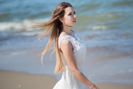 Young charming brunette woman on sea coast. Beautiful girl in a white summer dress. On the background of sea wavesの写真素材