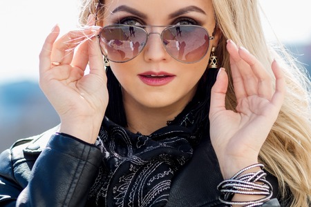 Close up image of beautiful young woman looking at camera. Young caucasian female model looking through sunglasses. Femme blonde in a leather jacketの写真素材