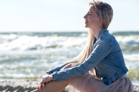 Young cheerful girl on the seashore. Young blonde woman sitting on the sand. Trendy beige skirtの写真素材