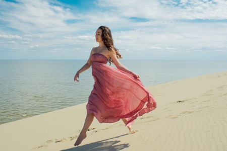 Woman in red waving dress with flying fabric runs on background of dunes. Skyline and the seaの写真素材