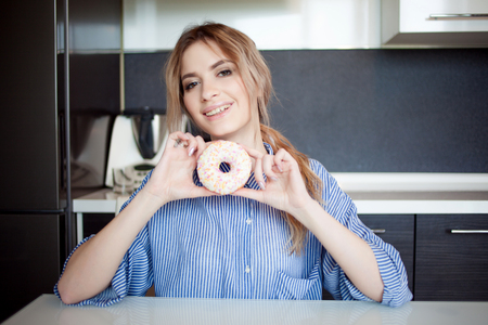 Beautiful young woman with a donut in hand. Girl on the kitchen tableの写真素材