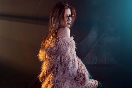 Young seductive woman, portrait in darkness. Girl in stylish pink fur coat and glasses. Smokey backgroundの写真素材