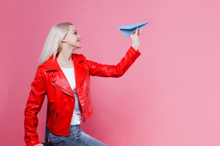 Happy young woman with travel suitcase launches paper airplane. Blonde tourist girl on pink background, concept, travel or vacationの写真素材