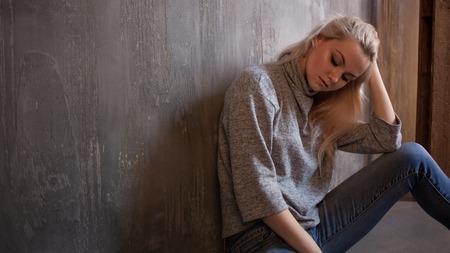 Depressing girl. sits on the floor. Depression and chronic fatigue. Young beautiful blonde in a gray sweater and jeans, gray textured backgroundの写真素材