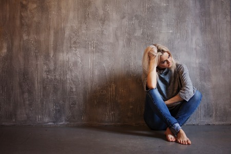 Sad woman sits on the floor. Depression and chronic fatigue. Young beautiful blonde in a gray sweater and jeans, gray textured backgroundの写真素材