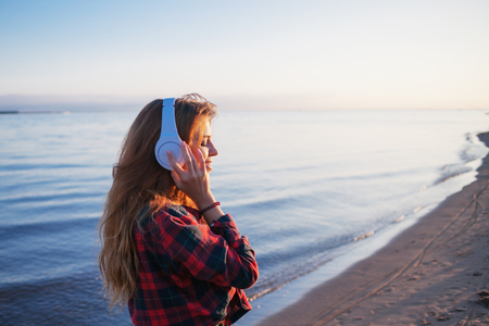 Happy attractive red-haired girl is enjoying favorite music on the walk. Beautiful young woman in shirt uses headphonesの写真素材