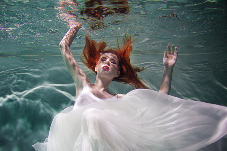Underwater girl. Beautiful red-haired woman in a white dress, swimming under water. Nymph or mermaid, fantasy concept.の写真素材