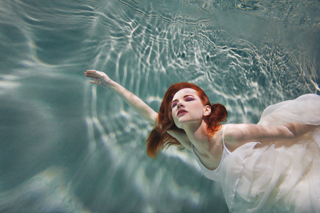 Underwater girl. Beautiful red-haired woman in a white dress, swimming under water. Nymph or mermaid, fantasy concept.の写真素材