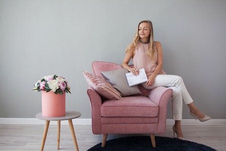Young friendly woman sitting in a chair with a notebook in her hands. Stylish interior design, a...