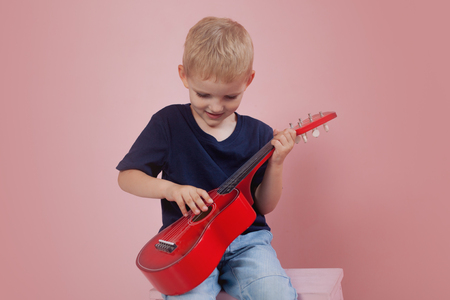 Little boy is learning to play on a small guitar. Study music. Ukulele. Portrait on pink backgroundの写真素材
