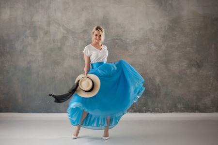 Beautiful young woman in a lush blue skirt, whirling and dancing. Summer style, straw hat with black ribbon. Grey textured backgroundの写真素材