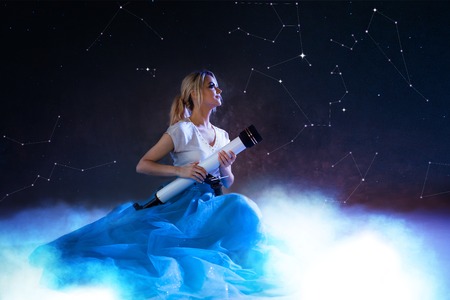 Young woman dreams of the future, concept. Girl in the sky with clouds looks up and uses a telescope. Dream and set goals, look through the telescope. Starry sky behind the cloudsの写真素材