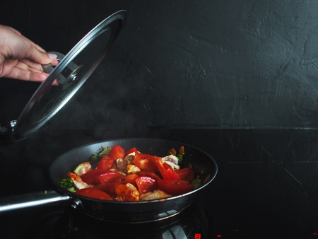 Frying pan with vegetables and chicken meat on an induction cooktop, closing cover, black background