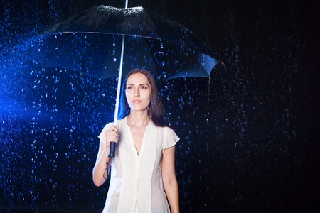 Young woman under umbrella. Protection from rain. Brunette girl holding an umbrella. Portrait in Studio on black background. copy space.の写真素材