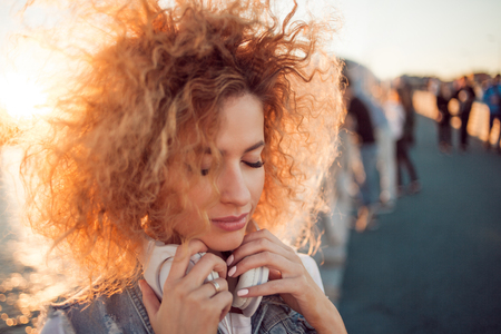 Trendy girl with large headphones on a city walk, close up. Portrait of a young charming blonde with lush curls, Enjoy the music and closed eyesの写真素材