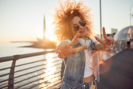 Young stylish woman with very lush hair show victory sign, focus on fingers. Young woman with big headphones. outdoors portrait of a trendy girlの写真素材