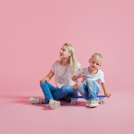 Positive young mother with a young son. Beautiful blonde with her baby on a skateboard, pink backgroundの写真素材