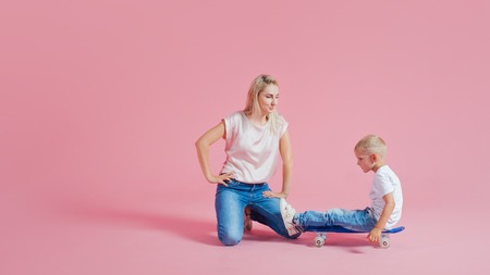 Positive young mother with a young son. Beautiful blonde with her baby on a skateboard, pink backgroundの写真素材