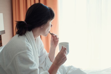 young woman in a white Terry robe woke up this morning. Drinking coffee in bed in the hotelの写真素材