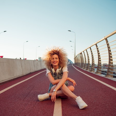 Happy young girl on the street. Beautiful blonde with headphones, Sitting on the bike pathの写真素材