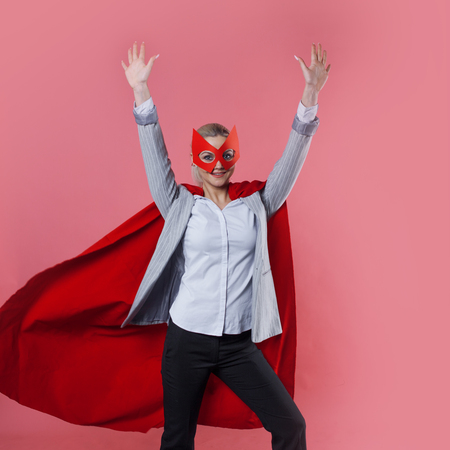 Young attractive woman superhero. Girl in a business suit and a mask with red cloak of hero. On a pink background.の写真素材