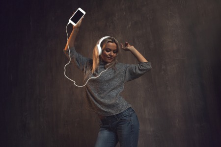 Girl dancing and listening to music in a mobile application. Neon white phone and earphone wire. Grey clothing and backgroundの写真素材